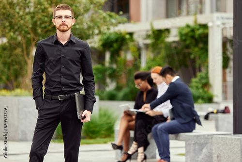Portrait of serious, concentrated young businessman in black clothes standing and looking at camera. Blurred employees on background