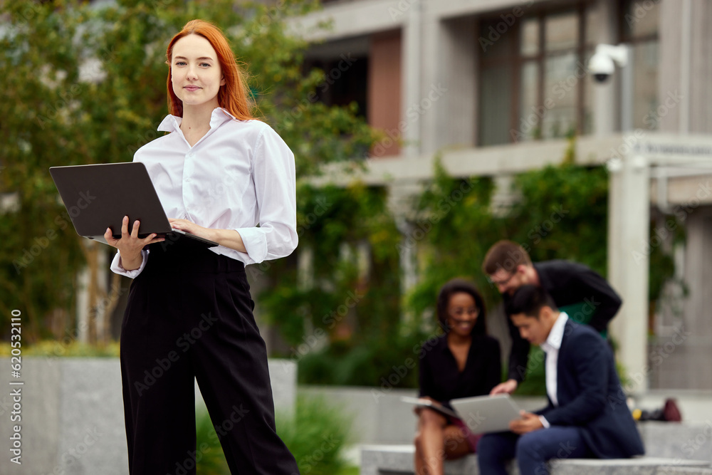 Fototapeta premium Portrait of serious, concentrated young woman, employee standing with laptop on street. Blurred employees on background