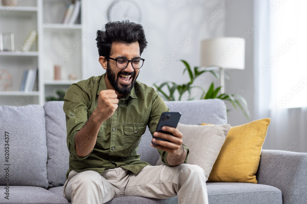 Happy and satisfied with achievement results hispanic at home reading online win notification in online application on phone, man smiling holding hand up triumph gesture sitting sofa in living room.