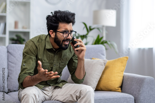 Photo Angry and nervous man talking on the phone sitting on the sofa in the living room, hispanic man yelling at the interlocutor on the smartphone, dissatisfied with the service, the customer complains