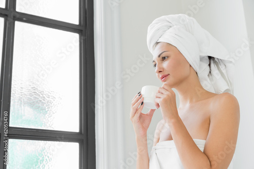 A beauty-loving woman wearing a towel with a healthy, smooth body, drinking coffee in a white bathtub in a bathroom with a large window.