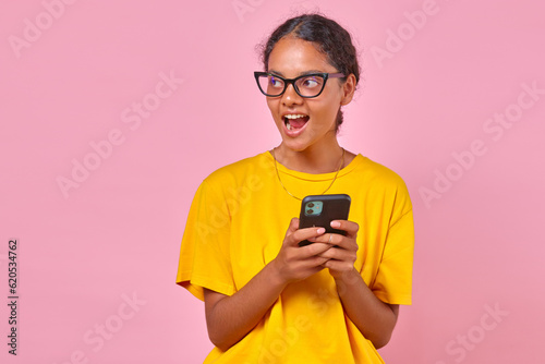 Young inspired Indian woman in casual clothes holding mobile phone and shouting loudly after reading alert in banking application about receipt of money in account stands on isolated pink background. © antianti
