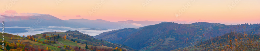 panorama of mountainous countryside at dawn. hills with trees and rural fields rolling down into the foggy valley. distant ridge with snow capped tops beneath a sky with clouds in red morning light