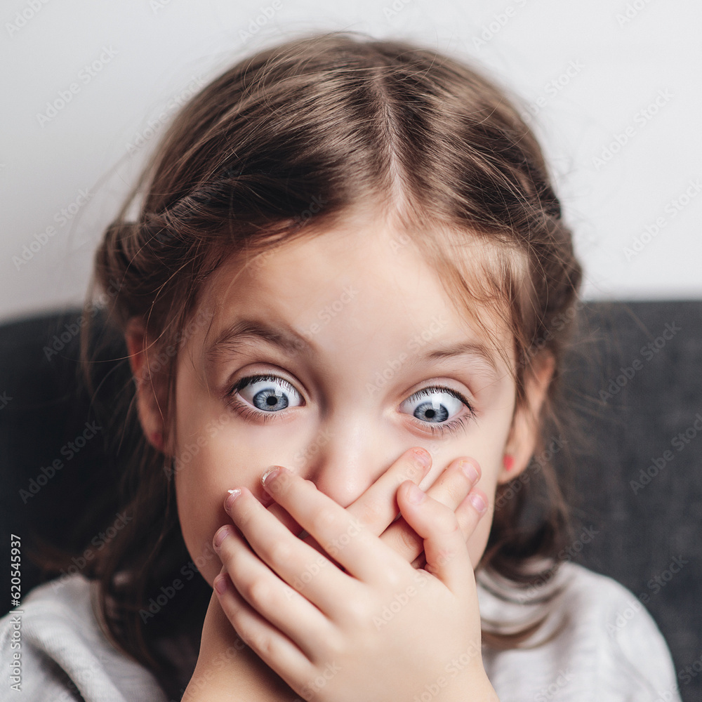 Little girl covers mouth with hand. Sad offended child girl sitting at sofa at home. Child abuse. Little girl covers mouth with hand. Sad offended child girl sitting at sofa at home. Child abuse.