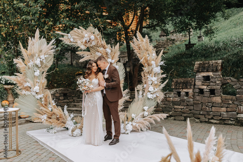 The bride and groom are enjoying themselves. Newlyweds with a wedding bouquet standing at a wedding ceremony under an arch decorated with flowers and dried flowers outdoors.