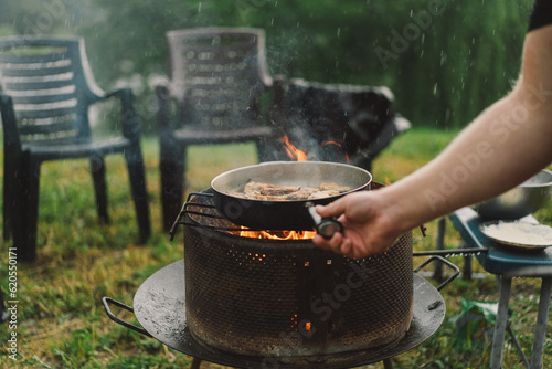 Cooking in the woods outdoors. Man cooking fish on fire in rainy weather, camping outdoors. Tourist on recreation outside. Campsite lifestyle