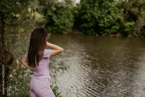 a girl in a lilac suit stands on the shore of a pond in summer. a walk in the park.