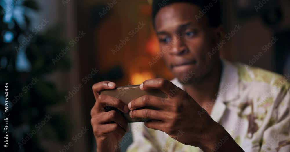 African man takes break and sits on couch, engrossed in mobile gaming on his cellphone experiences disappointment struggles to conquer difficult level, but remains determined to improve succeed. African man takes break and sits on couch, engrossed in mobile gaming on his cellphone experiences disappointment struggles to conquer difficult level, but remains determined to improve succeed.
