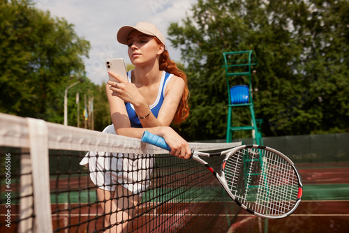 Active, fit and happy female tennis player browsing social media on her phone outdoors on the court. A young female athlete or sportswoman posting her sport training online or on the internet © st.kolesnikov