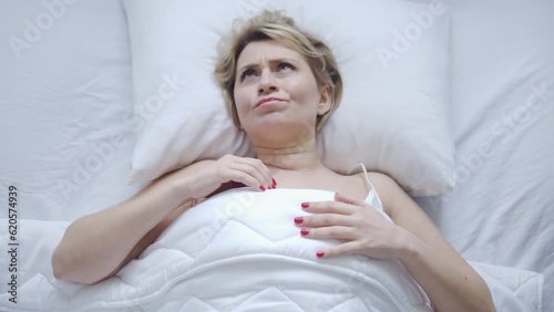 Unhappy woman lying in bed feeling pain and discomfort in her head, neck and shoulders because of uncomfortable cheap pillow, bad sleeping conditions, lack of orthopedic bedding photo