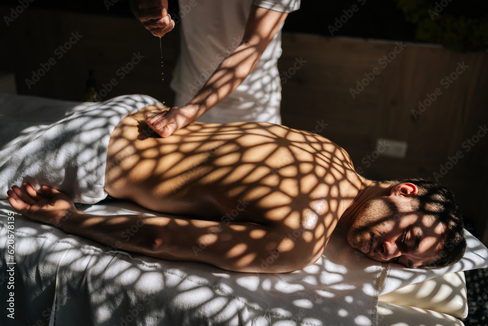 Obraz premium Relaxed young man getting massage in spa salon lying with closed eyes on massage table in sunlight and beautiful shadows. Closeup hands of massage therapist stroking back of client in wellness center.