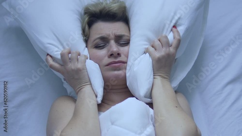 Woman crying and covering ears with pillow, irritated with noise sound of repairs or loud party at the neighbors, working alarm or thunder and rain outside the window