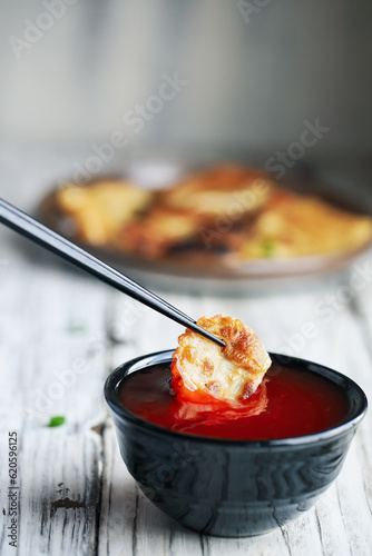 Chopsticks dipping a golden fried Chinese dumpling, also known as Pot Sticker, into a bowl of red sweet and sour sauce.  Selective focus with blurred background.