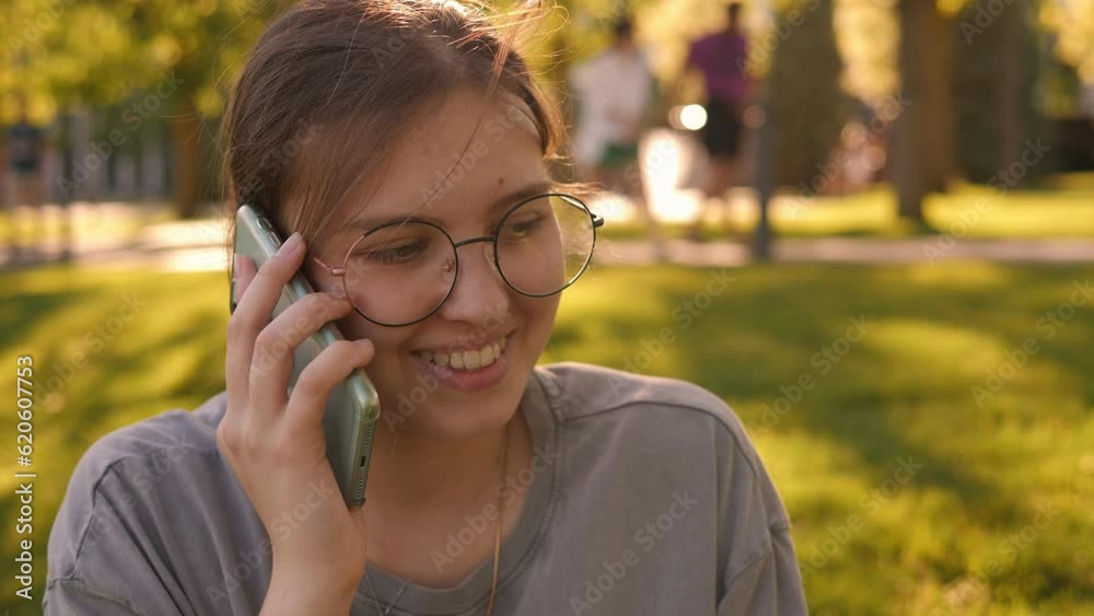 Close-up of a teenage girl talking on the phone in the park while sitting on a grass in the summer at sunset. Close-up of a teenage girl talking on the phone in the park while sitting on a grass in the summer at sunset.