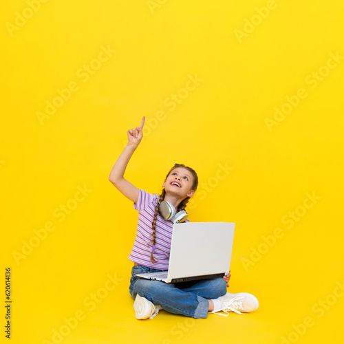 A child with a laptop. A little girl sits on the floor and communicates online on a laptop, wearing headphones. Internet communication for children. Yellow isolated background. copy space. © Юлия Дьякова