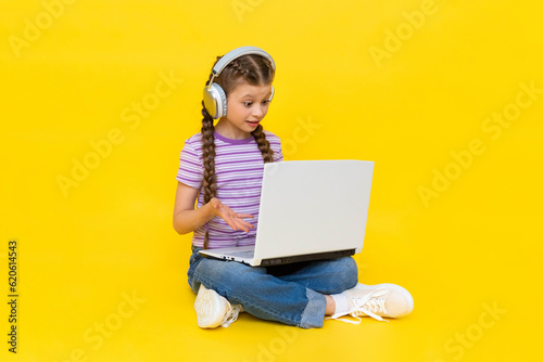 Internet communication for children. A young girl with a laptop in her hands is sitting on the floor. The child communicates on social networks online. Yellow isolated background.