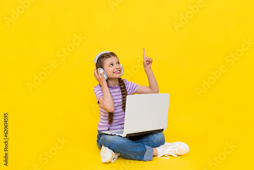 A child with a laptop. A little girl sits on the floor and communicates online on a laptop, wearing headphones. Internet communication for children. Yellow isolated background. copy space.