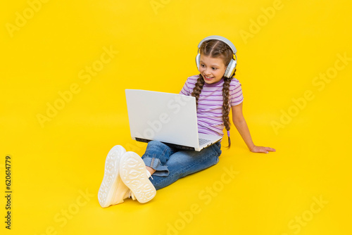 Internet communication for children. A young girl with a laptop in her hands is sitting on the floor. The child communicates on social networks online. Yellow isolated background.