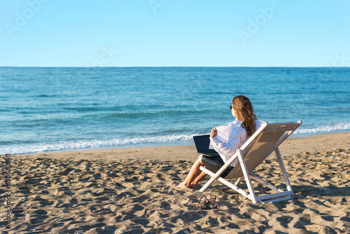 Back view of alone young brunette woman freelancer in office clothes with a laptop looking on the sea sitting on the deck chair on beach. Summer concept