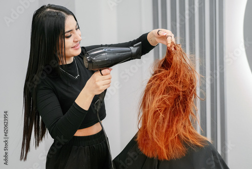 Woman hairdresser does hair styling with hairdryer to client in beauty salon in front of mirror. Woman with red hair sits in chair in hairdresser. Professional stylist makes hairstyle.