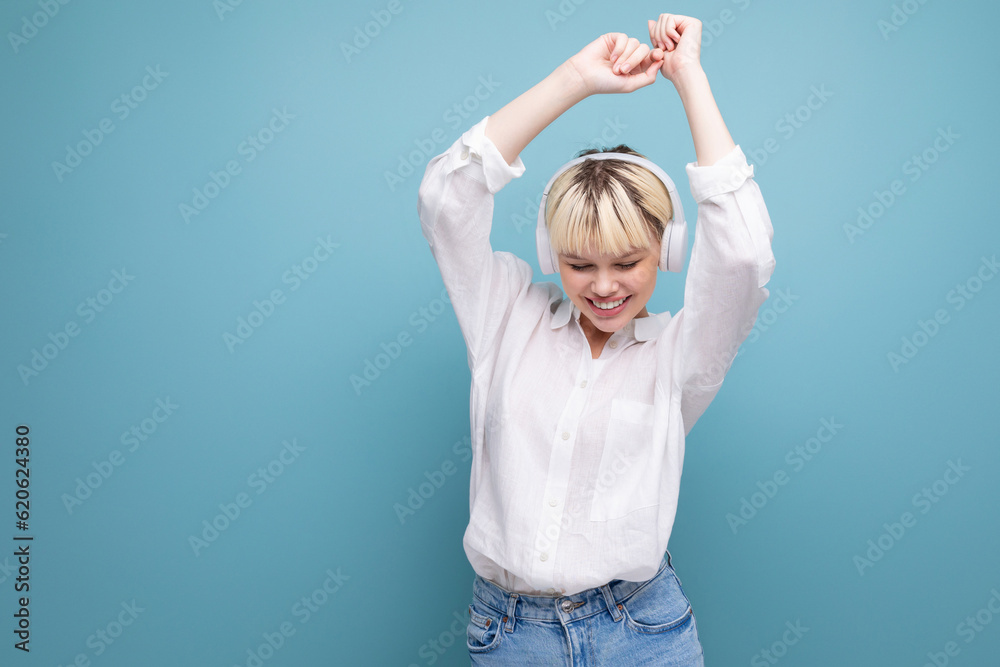 Fototapeta premium young pretty blond student woman dressed in a white blouse listens to her favorite music in headphones without a wire on the background with copy space