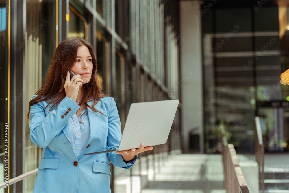 Beautiful businesswoman wearing blue jacket using laptop and talking on mobile phone outdoors