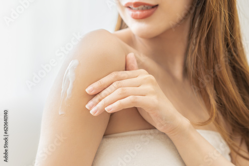 Health care  beauty smile asian young woman  girl wrapped in towel after shower bath  spa relax at home  hand applying  putting moisturizing lotion on her shoulder. Skincare routine in the morning.