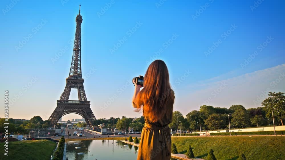 A beautiful woman in a dress uses a camera to picture the romantic city of Paris, France. Female tourist taking pictures of the historical Tower and enjoying the view of a golden sunset.