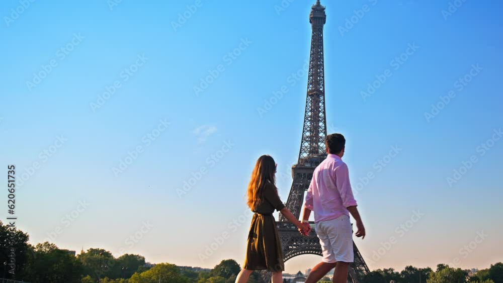 A young couple enjoys the view of Paris pointing at the Tower. A romantic woman and her boyfriend walk together holding hands at golden sunset in Paris, France.