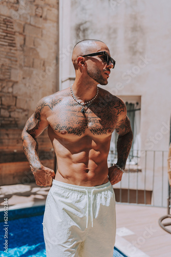 Chico joven musculado y tatuado posando  sin camiseta en piscina de estilo  boho en un hotel un dia soleado de verano photo