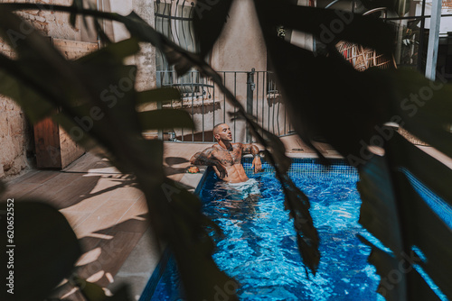 Chico joven musculado y tatuado posando sin camiseta en piscina de estilo boho en un hotel un dia soleado de verano