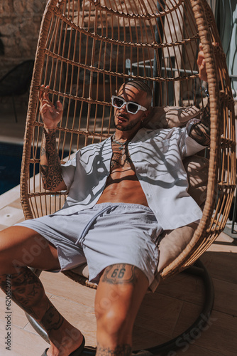 Chico joven tatuado posando con ropa de verano actual  en piscina estilo boho en un hotel un día de verano