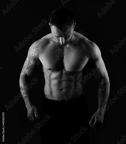 Strong man with bare muscular torso, studio black and white photo on black background. Bodybuilder strains body muscles.