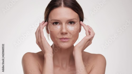 Portrait of healthy woman putting eye patche, touching her face while looking to camera and smiling. Beauty treatment. Caucasian Female model with naked shoulders and nude makeup posing.
