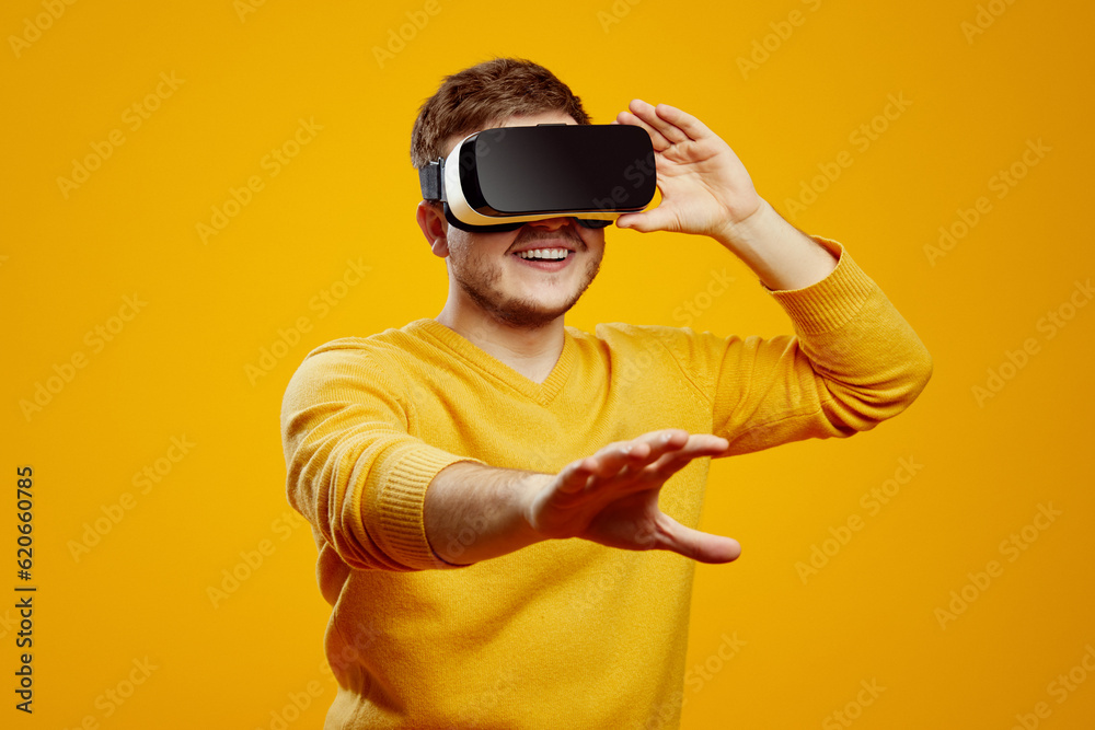 Young man in yellow sweater posing isolated on orange background. Watching in headset, touching floating virtual screen Young man in yellow sweater posing isolated on orange background. Watching in headset, touching floating virtual screen