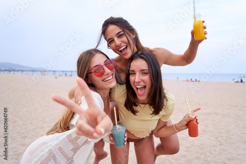 Cheerful portrait of three women on the beach playing doing piggyback while looking at camera smiling and gesturing the peace sing. Female friends with smoothies in summertime having fun.