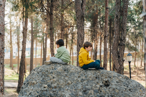Billede på lærred School kids boys having a conflict quarrel sitting on a rock boulder in the city forest park and turned their backs to each other