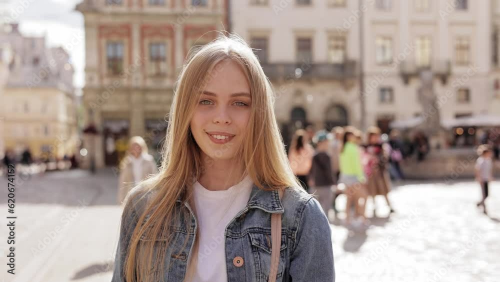 Close up portrait of pretty young blond female standing on background of crowded street and looking at camera smiling. Smiling Beautiful Young Girl Loking into Camera and Smiling at City Background. Close up portrait of pretty young blond female standing on background of crowded street and looking at camera smiling. Smiling Beautiful Young Girl Loking into Camera and Smiling at City Background.