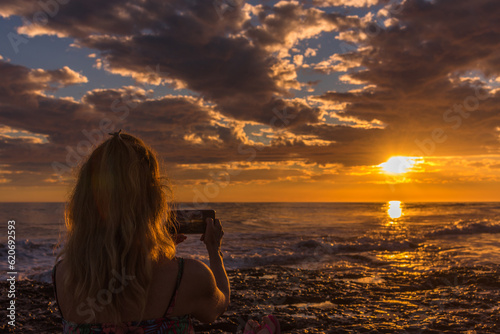Woman taking photos of the sunset on the beach with mobile phone. Summer, travel and technology concept.