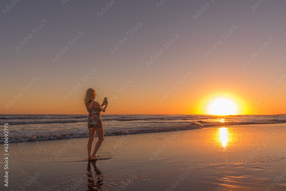 Woman with mobile phone taking photos of sunset standing on the seashore at the beach. Summer, travel and concept.