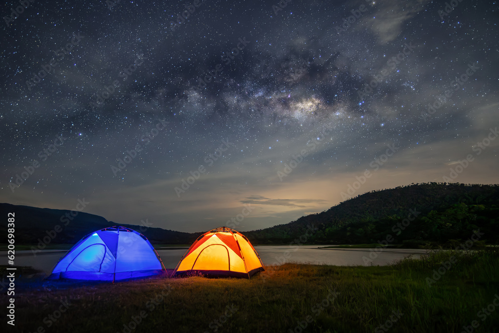 Panorama of the night sky with camping tents and lakes, surrounded by mountains. Milky way and stars on dark background with noise and grain. Long exposure shot with white balance selected.