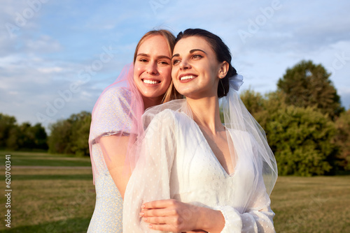 Happy lovely bride in white dress with white veil and bridesmaid in blue dress with pink veil in summer park. Young charming girls smile and hug. Cheerful bachelorette party. Close-up portrait. © Georgii