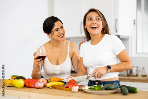 Happy female friends with glasses of red wine chatting and preparing salad together in modern kitchen
