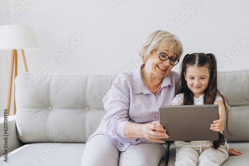 Family, generation, technology - smiling granddaughter and grandmother with tablet pc computer sitting on couch at home. Cheerful Girl sitting at home with her grandmother using tablet computer photo