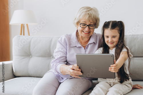 Family, generation, technology - smiling granddaughter and grandmother with tablet pc computer sitting on couch at home. Cheerful Girl sitting at home with her grandmother using tablet computer
