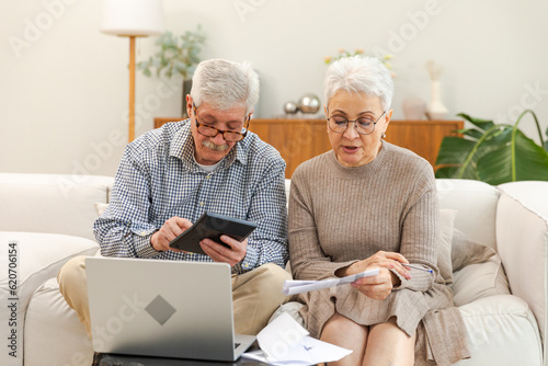 Middle aged senior couple sit with laptop and paper document. Older mature man woman reading paper bill pay online at home managing bank finances calculating taxes planning loan debt pension payment