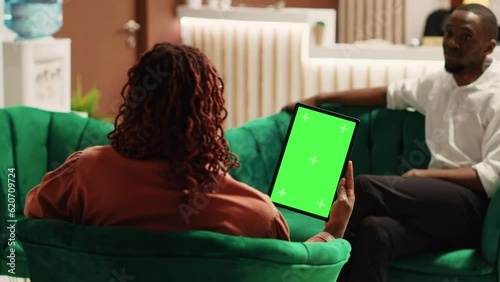 Happy guests in hotel lounge holding chroma key green screen mock up tablet while enjoying cup of coffee. African american couple waiting to be checked in, sitting on cozy sofa
