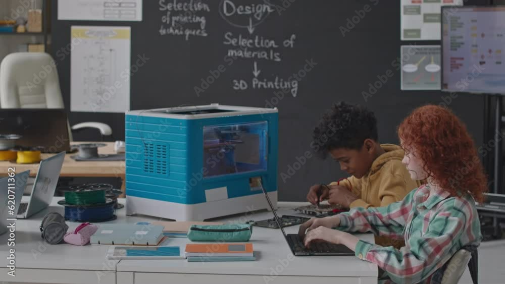 Little red haired girl using laptop and talking with African American classmate repairing computer detail with soldering iron at desk during 3D printing lesson
