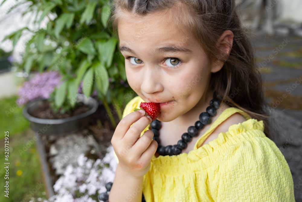A girl in yellow sits near a flower bed in homemade blueberry beads and eats ripe strawberries A girl in yellow sits near a flower bed in homemade blueberry beads and eats ripe strawberries