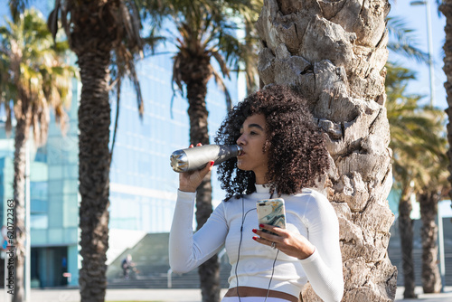 Ethnic sportswoman drinking water and listening to music in earp photo